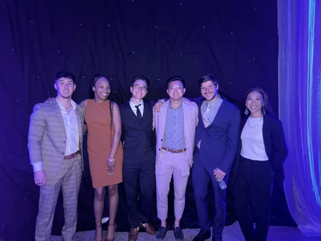 Group of six individuals posing together at a social event with a dark backdrop.