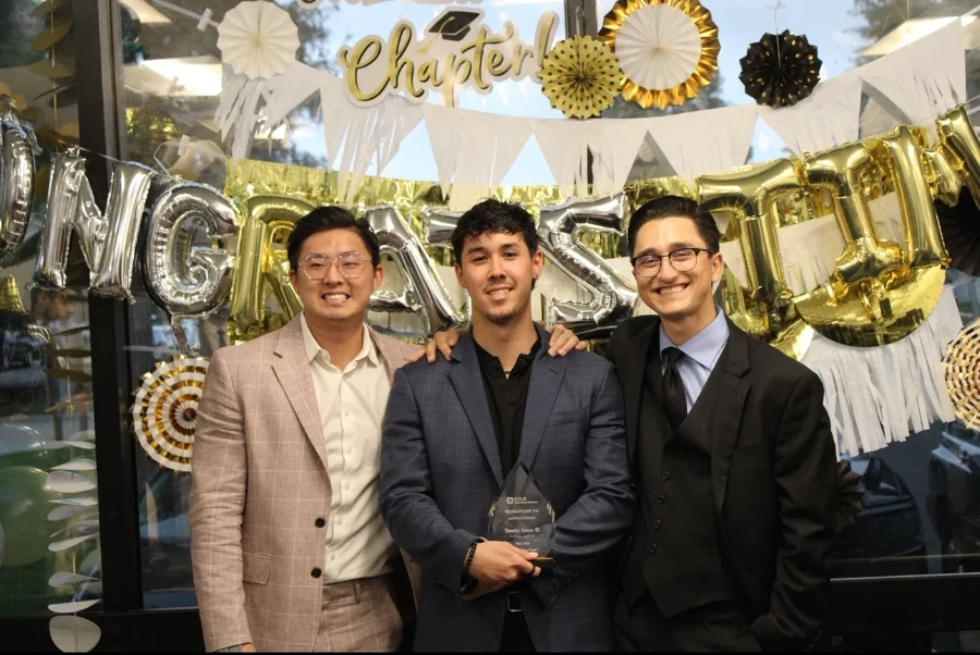 Three men posing together at a celebration, holding an award, with festive decorations in the background.